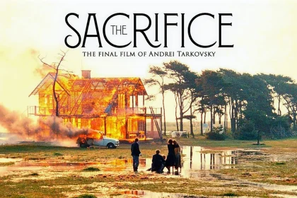 Film afişinde “SACRIFICE” yazısı üstte yer alır; altta açık arazide bir ev alevler içinde yanar, duman yükselir. Ön planda su birikintileri olan zeminde birkaç kişi yangına doğru bakar; sağ tarafta ağaçlar ve açık gökyüzü görülür.