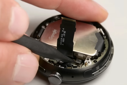 Google Pixel Watch’un açılmış kasası içinde pil ve bağlantı şeridinin yakın plan görüntüsü; bir el, plastik aparatla iç bileşenleri ayırıyor.